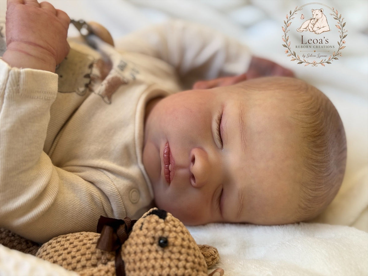 A reborn baby in a white ribbed romper asleep on a blue blanket, with a dummy on a beaded clip