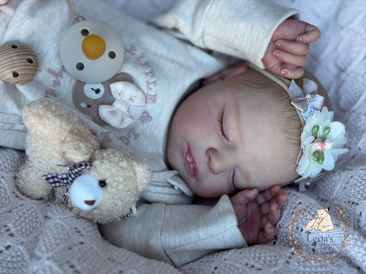 A reborn baby in a floral headband asleep on a cream blanket